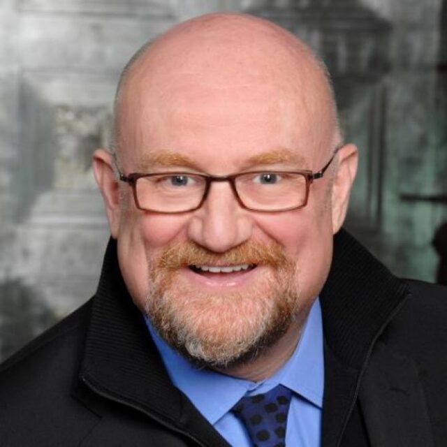 Portrait of a bald man with glasses and a ginger beard, smiling, wearing a blue shirt and black jacket