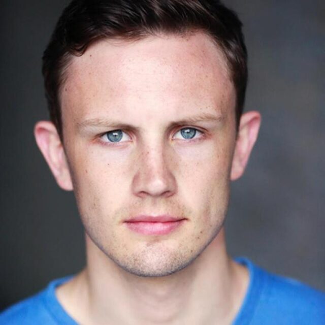 Headshot of a young man with short brown hair and blue eyes, wearing a blue top, against a grey background