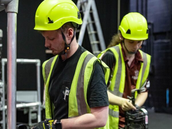 Two crew members in hard hats and hi-vis vests handling equipment amid scaffolding and rigging backstage