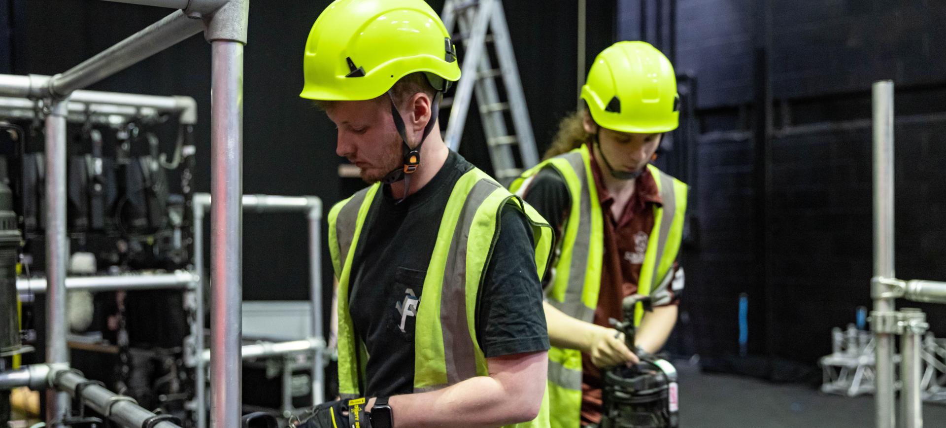 Two crew members in hard hats and hi-vis vests handling equipment amid scaffolding and rigging backstage