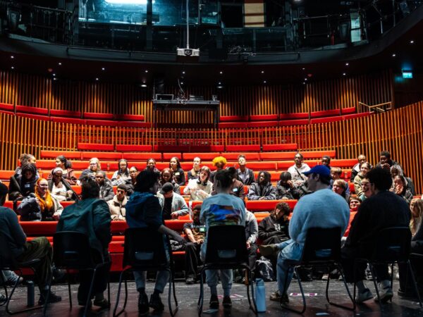 A large group sits in a circle on a theatre stage, some in chairs and others on tiered red auditorium seating, in discussion