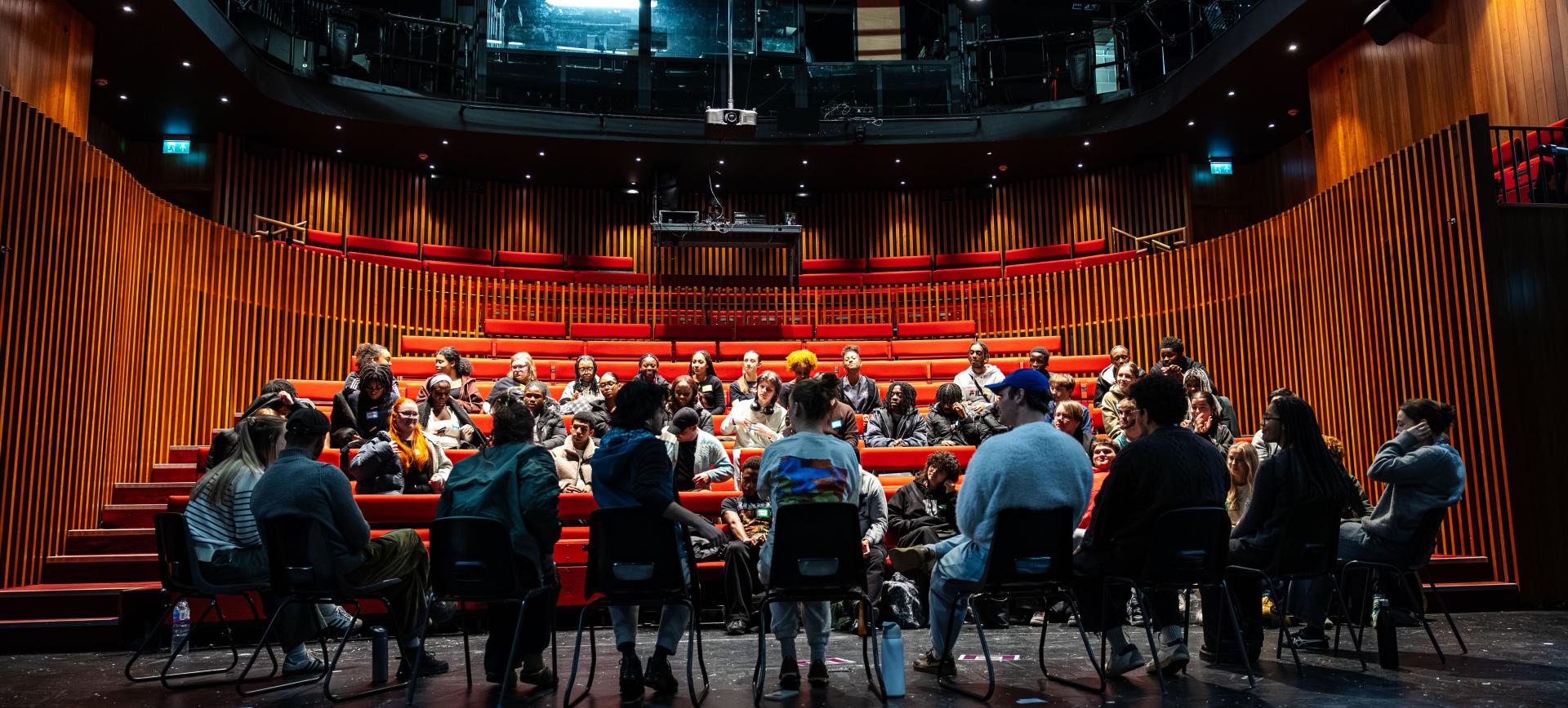 A large group sits in a circle on a theatre stage, some in chairs and others on tiered red auditorium seating, in discussion