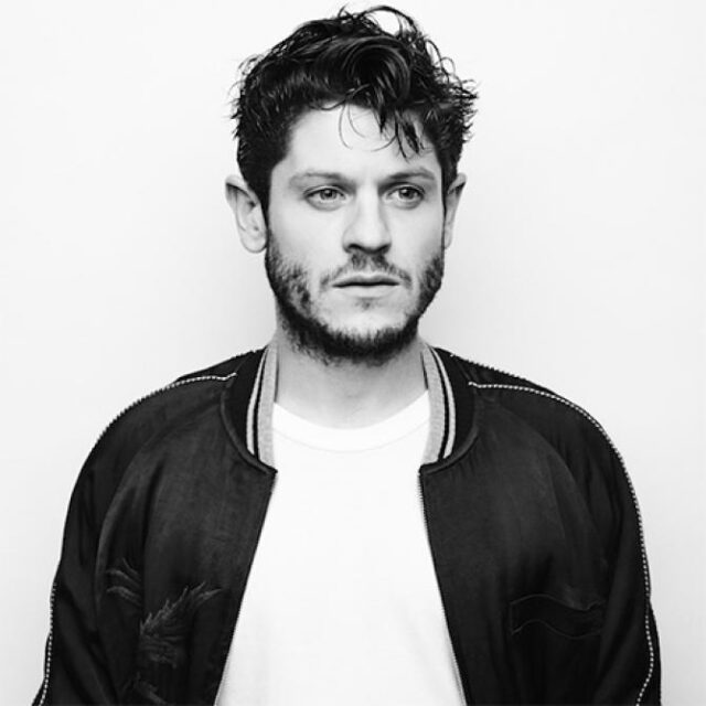 Black-and-white portrait of a man with tousled hair and beard, wearing a bomber jacket over a white T-shirt