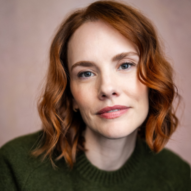 Woman with wavy red hair wearing a dark green jumper, looking directly at the camera