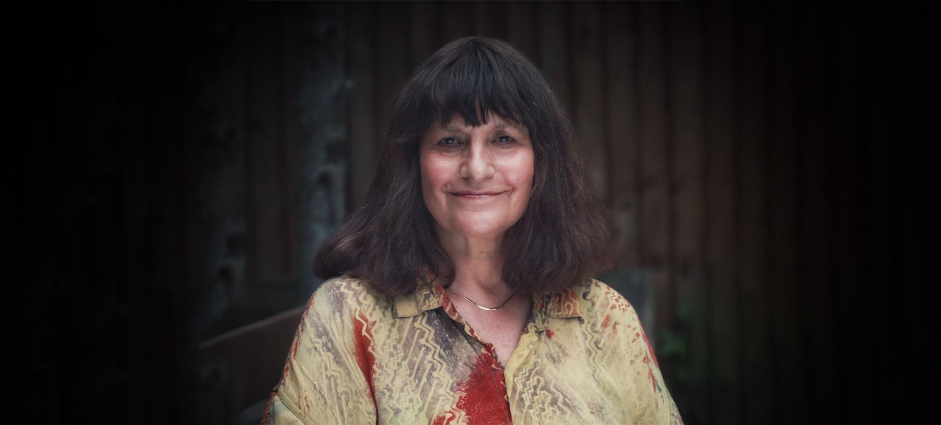Woman with dark shoulder-length hair and a fringe, smiling, wearing a patterned red and gold shirt
