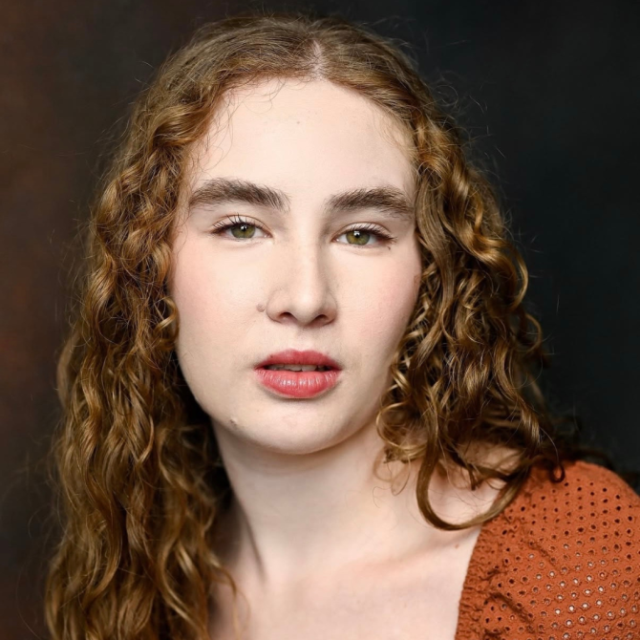 Portrait of a young woman with long curly reddish-blonde hair, wearing a rust-coloured top