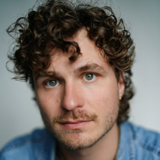 Close-up portrait of a man with curly brown hair, blue eyes and light stubble, wearing a denim shirt