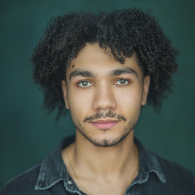 Portrait of a young man with curly dark hair and light eyes, wearing a dark denim shirt, against a dark green background