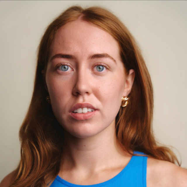 Close-up portrait of a young woman with long red hair and blue eyes, wearing gold hoop earrings and a blue top