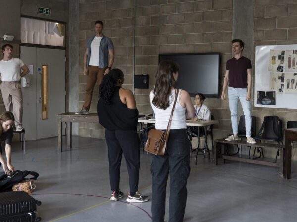 Actors stand on tables and chairs during a rehearsal, while others watch and gather bags in a studio room