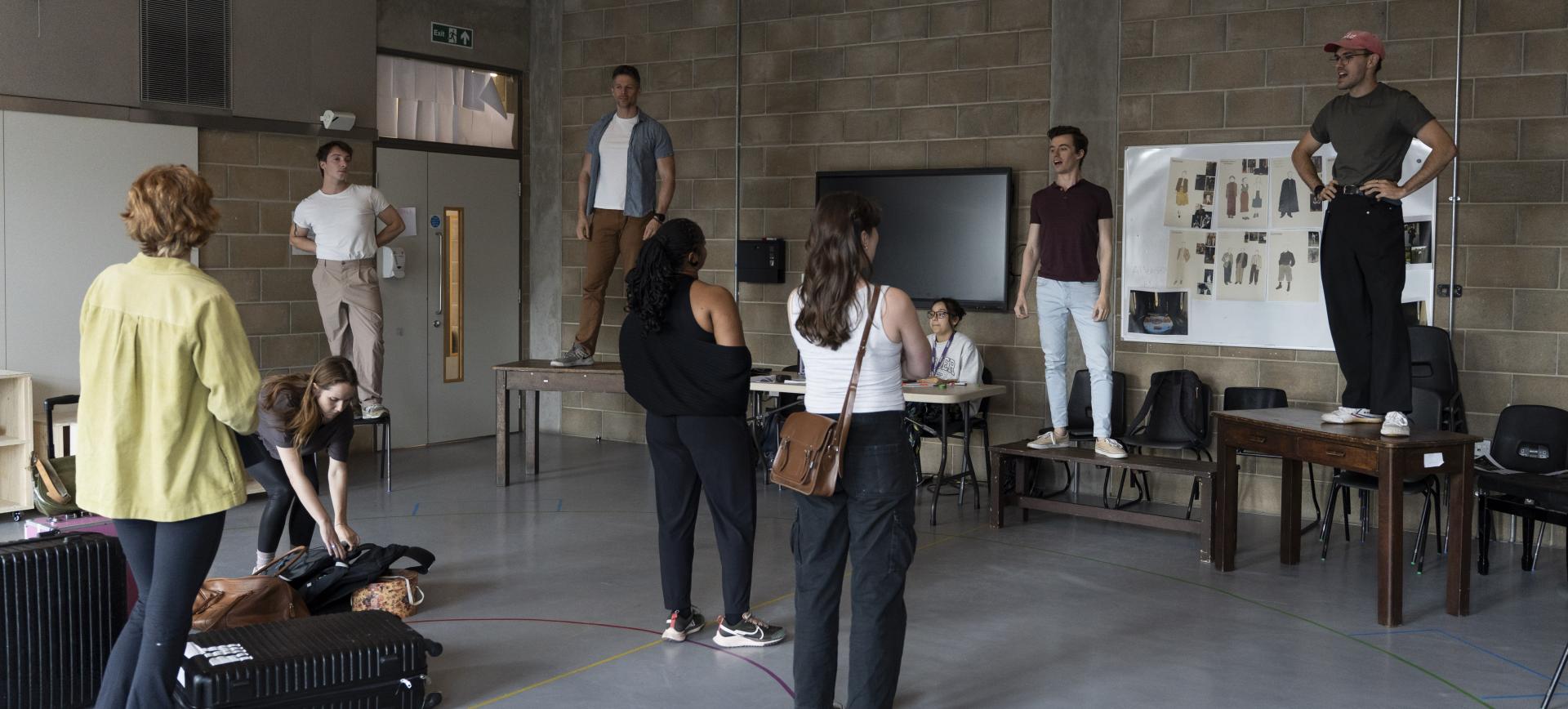 Actors stand on tables and chairs during a rehearsal, while others watch and gather bags in a studio room