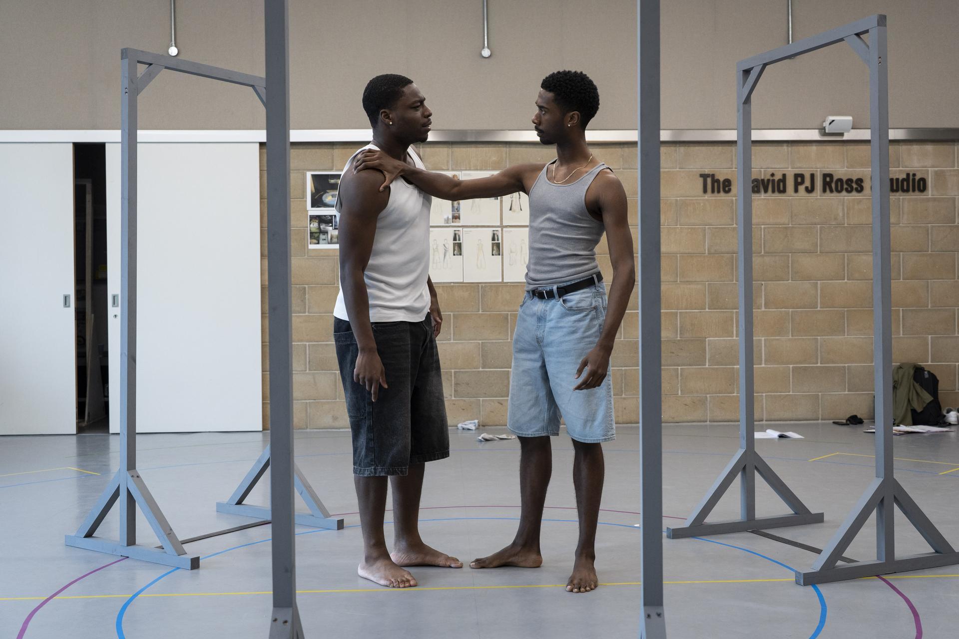 Two actors rehearse barefoot in a studio, one resting a hand on the other's shoulder in a tense exchange