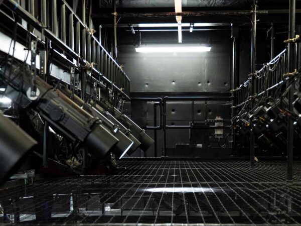 Rows of stage lighting fixtures mounted on rigging above a metal grid catwalk in a dark theatre fly system