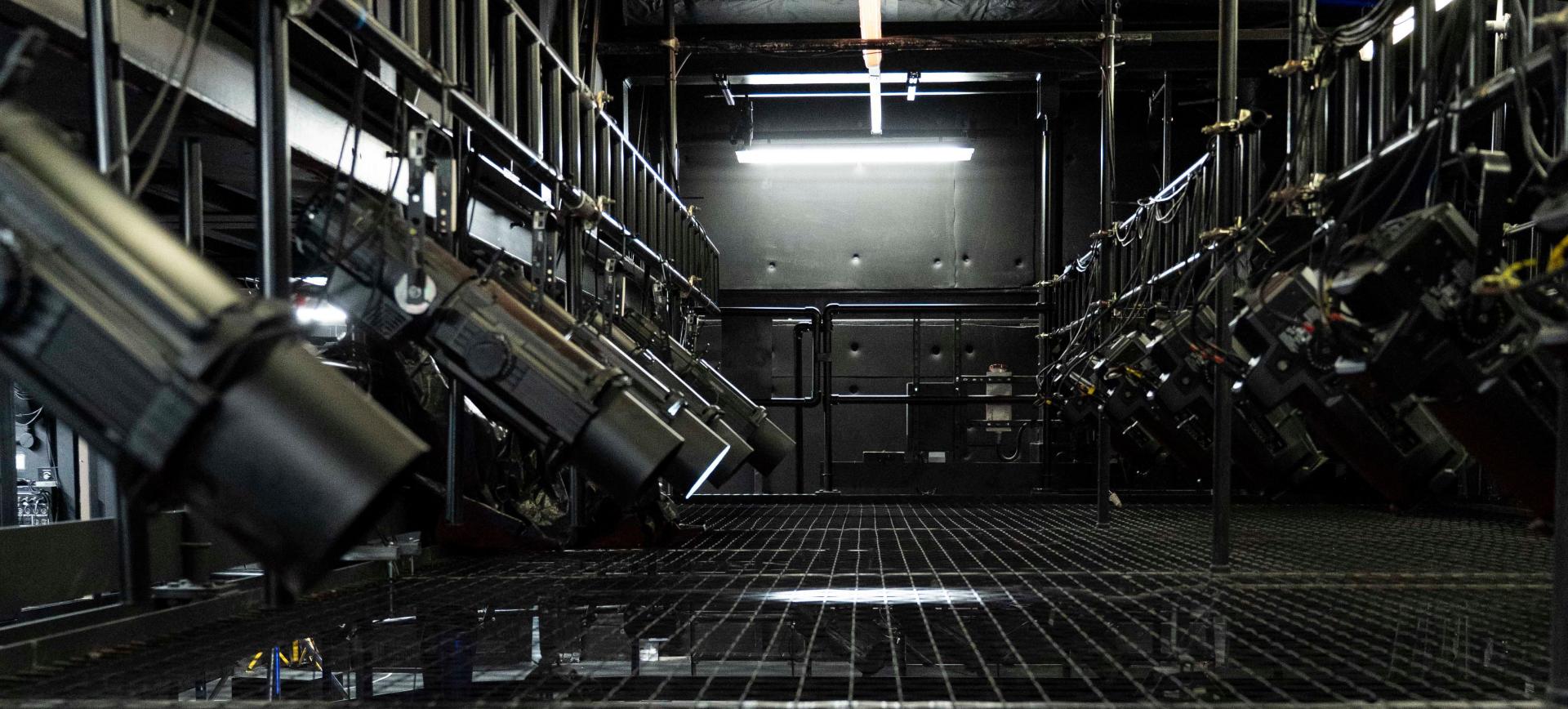 Rows of stage lighting fixtures mounted on rigging above a metal grid catwalk in a dark theatre fly system