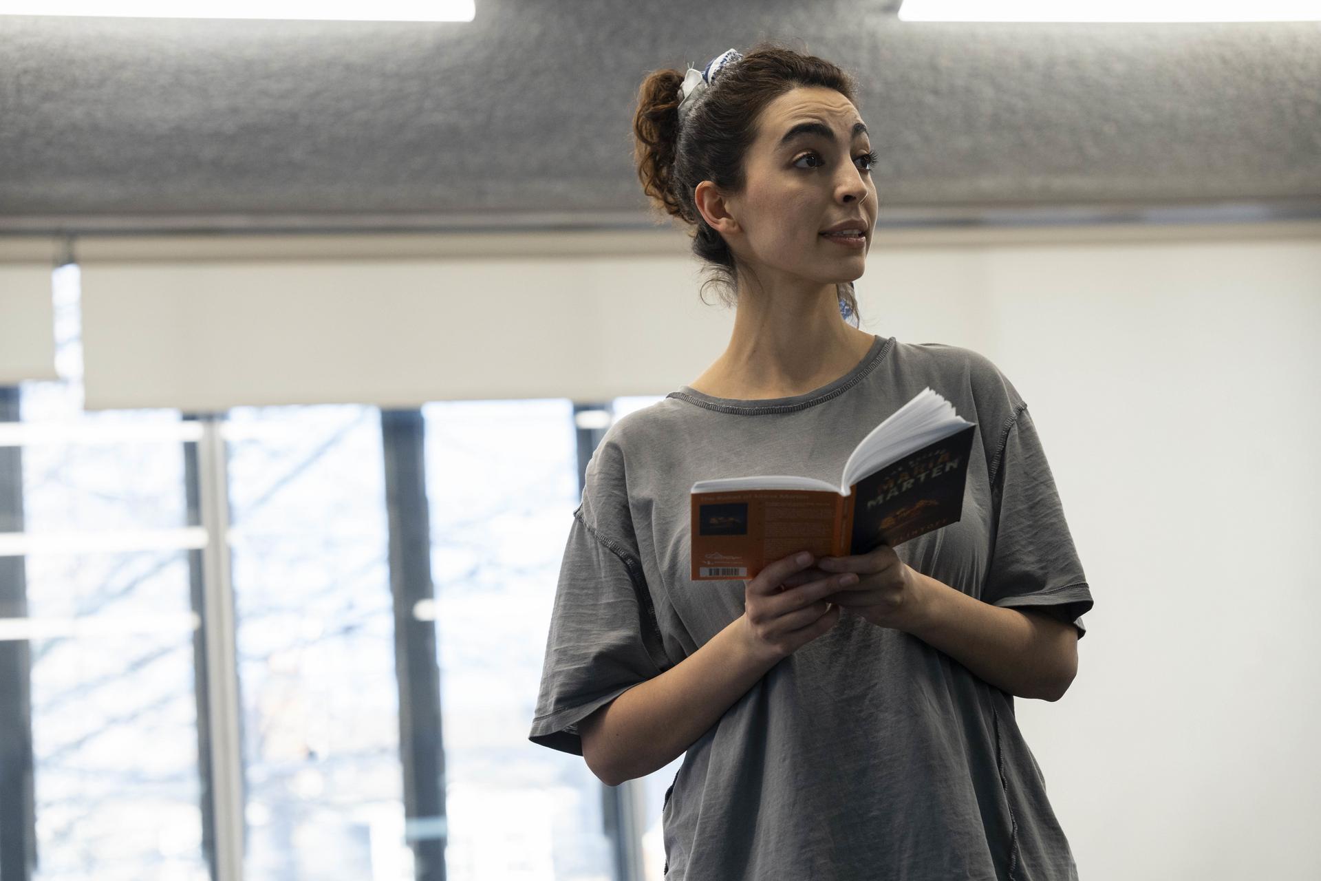 A woman rehearses a scene while holding an open copy of "Maria Marten", looking off to the side