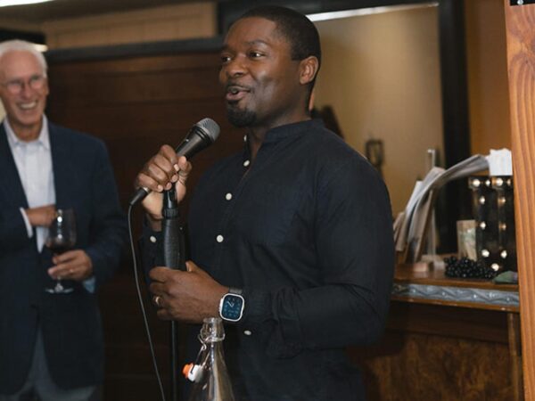 Man speaking into a microphone at an event, with another man laughing while holding a glass of wine nearby