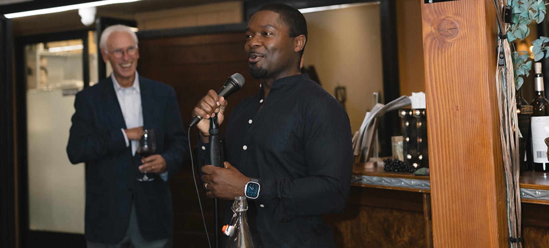 Man speaking into a microphone at an event, with another man laughing while holding a glass of wine nearby