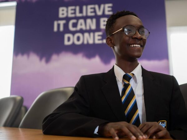 Smiling schoolboy in blazer and tie sits at a table, with a "Believe it can be done" banner behind him