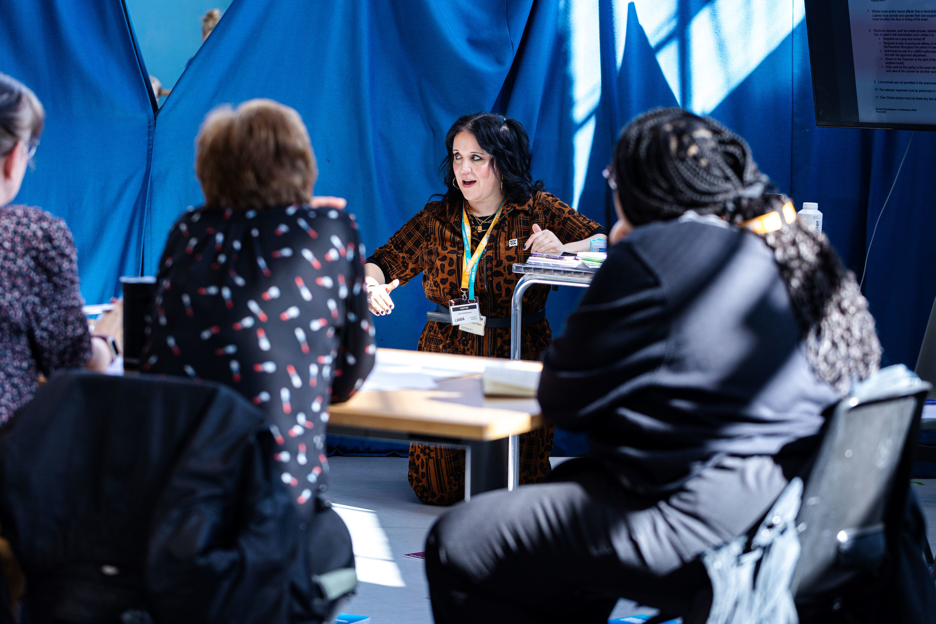 A LAMDA visitor wearing a lanyard kneels while speaking to a group seated around a table in a curtained booth