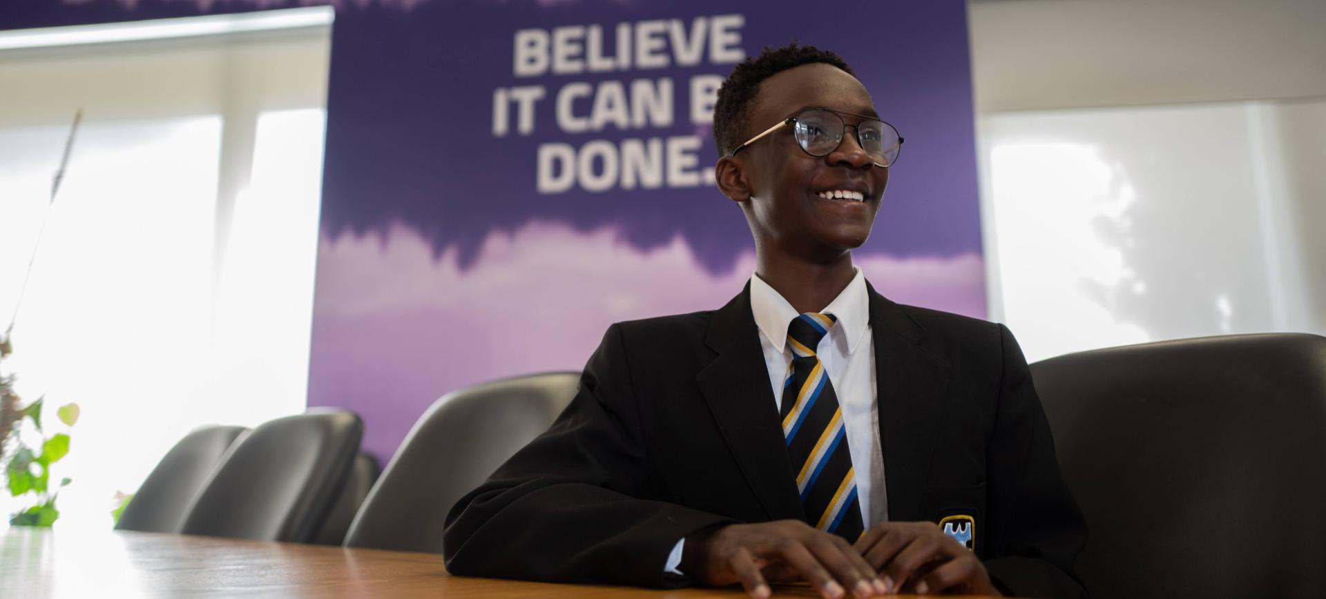 Smiling schoolboy in blazer and tie sits at a table, with a "Believe it can be done" banner behind him