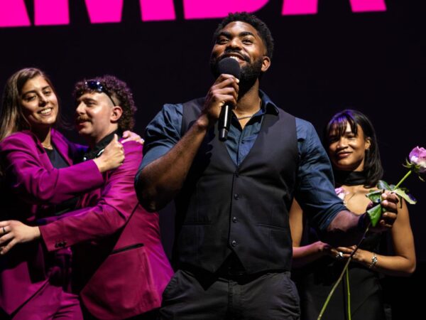Man speaking into a microphone on stage, smiling, with three castmates behind him, one holding a rose