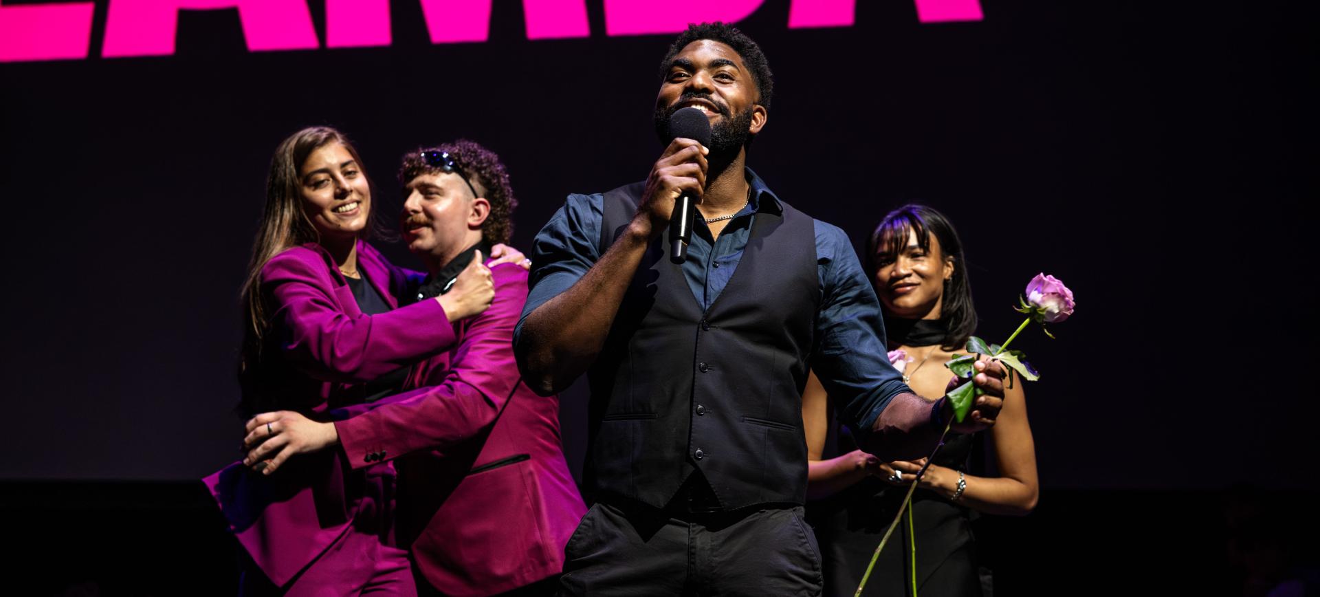 Man speaking into a microphone on stage, smiling, with three castmates behind him, one holding a rose