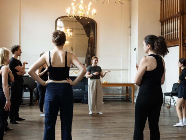 Dancers in rehearsal wear gather in a circle in an ornate studio, listening to an instructor speaking