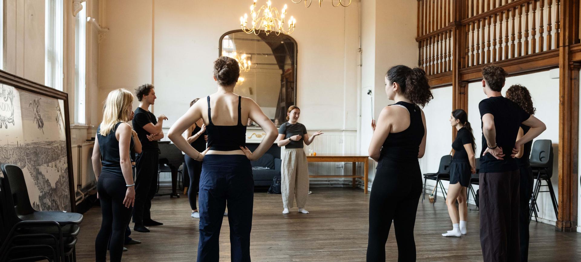 Dancers in rehearsal wear gather in a circle in an ornate studio, listening to an instructor speaking