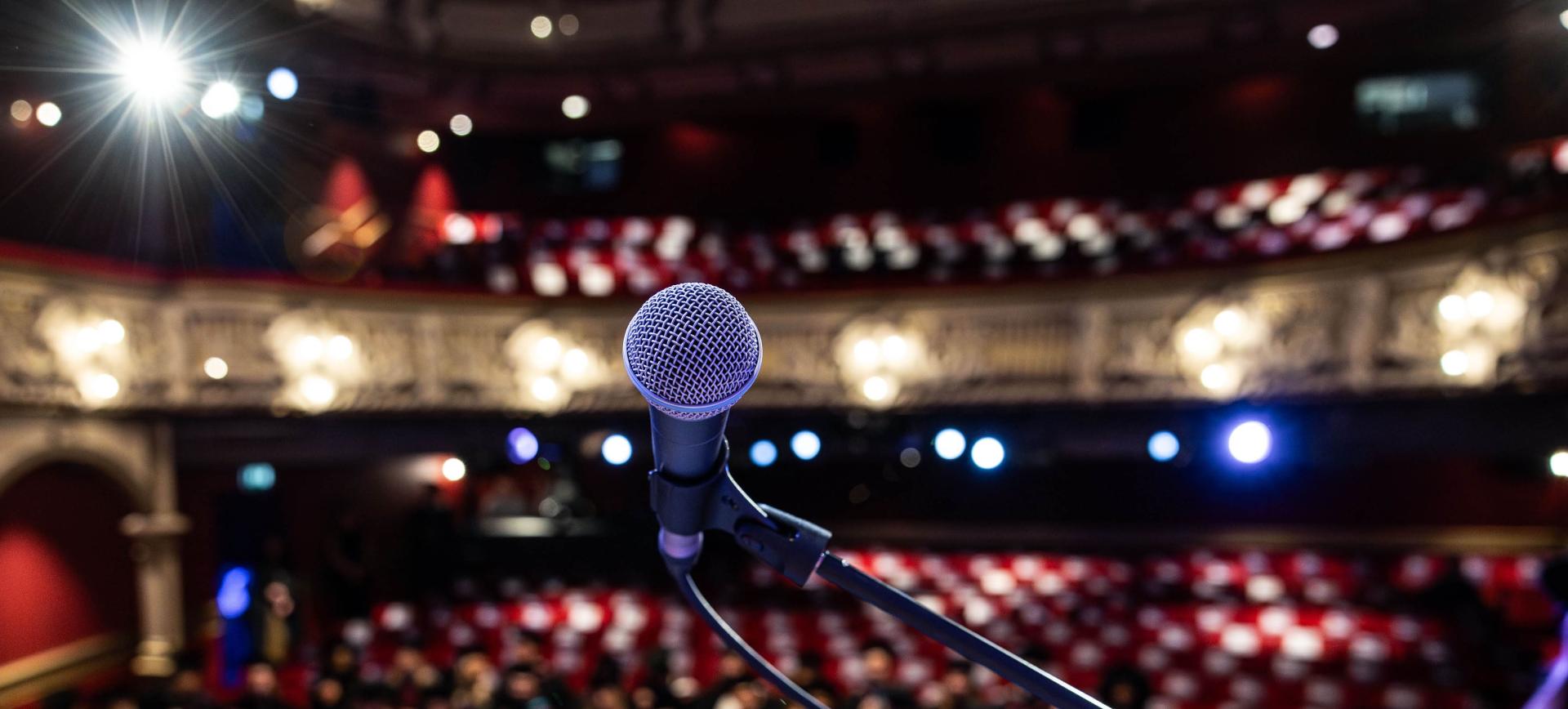 Close-up of a microphone on stage facing an empty theatre auditorium with red seating and stage lights