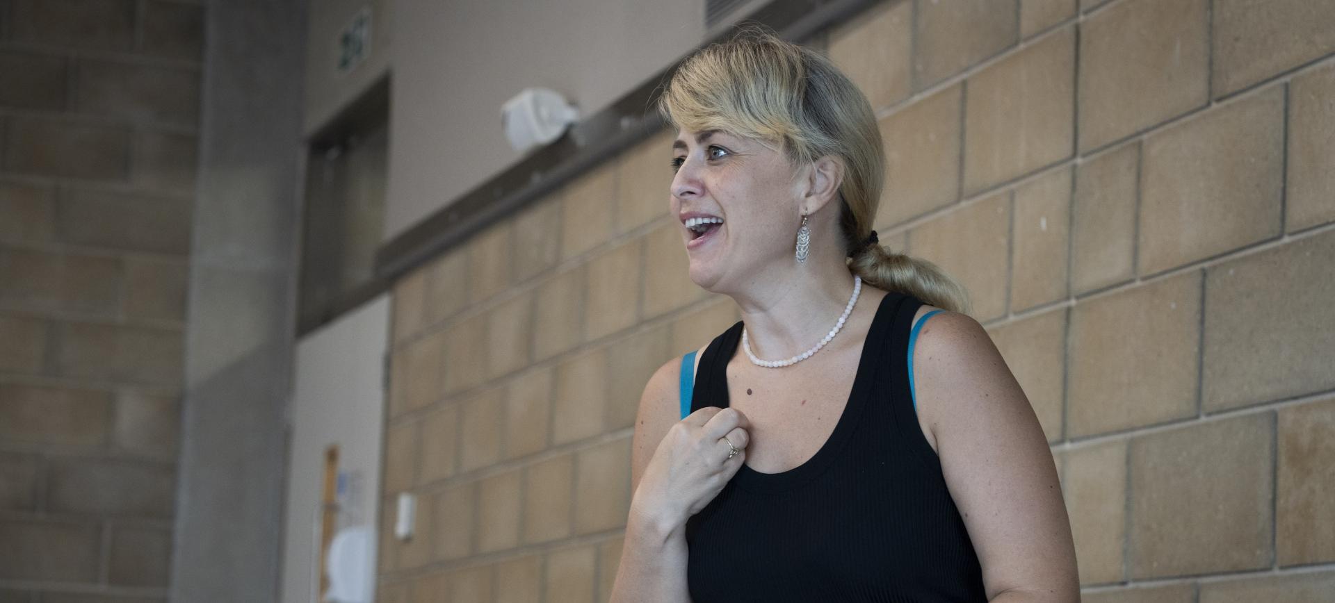 Woman with blonde hair and a pearl necklace smiling and talking, wearing a black vest top