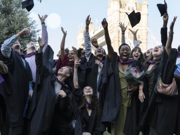 Graduates in gowns throw their caps in the air near Tower Bridge, holding LAMDA Certificate of Graduation folders