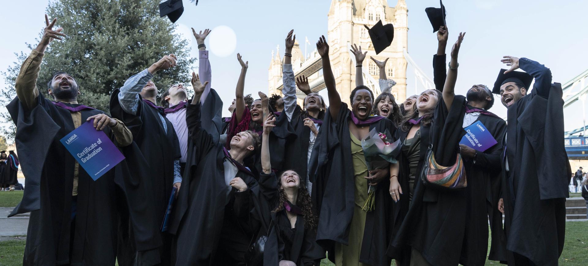 Graduates in gowns throw their caps in the air near Tower Bridge, holding LAMDA Certificate of Graduation folders