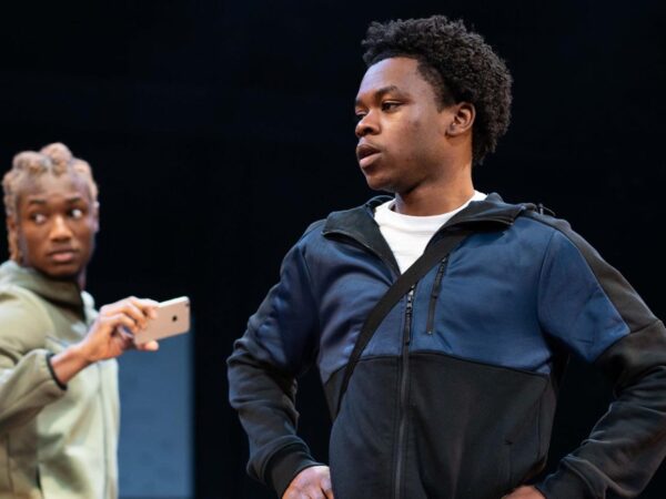 Actor stands with hands on hips as another holds up a phone to film him on a dark stage