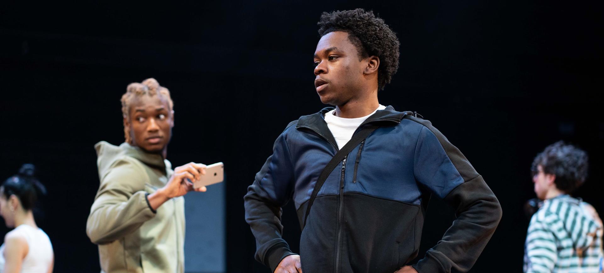 Actor stands with hands on hips as another holds up a phone to film him on a dark stage