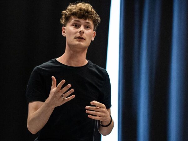 Young man with curly hair speaking on stage, gesturing with both hands