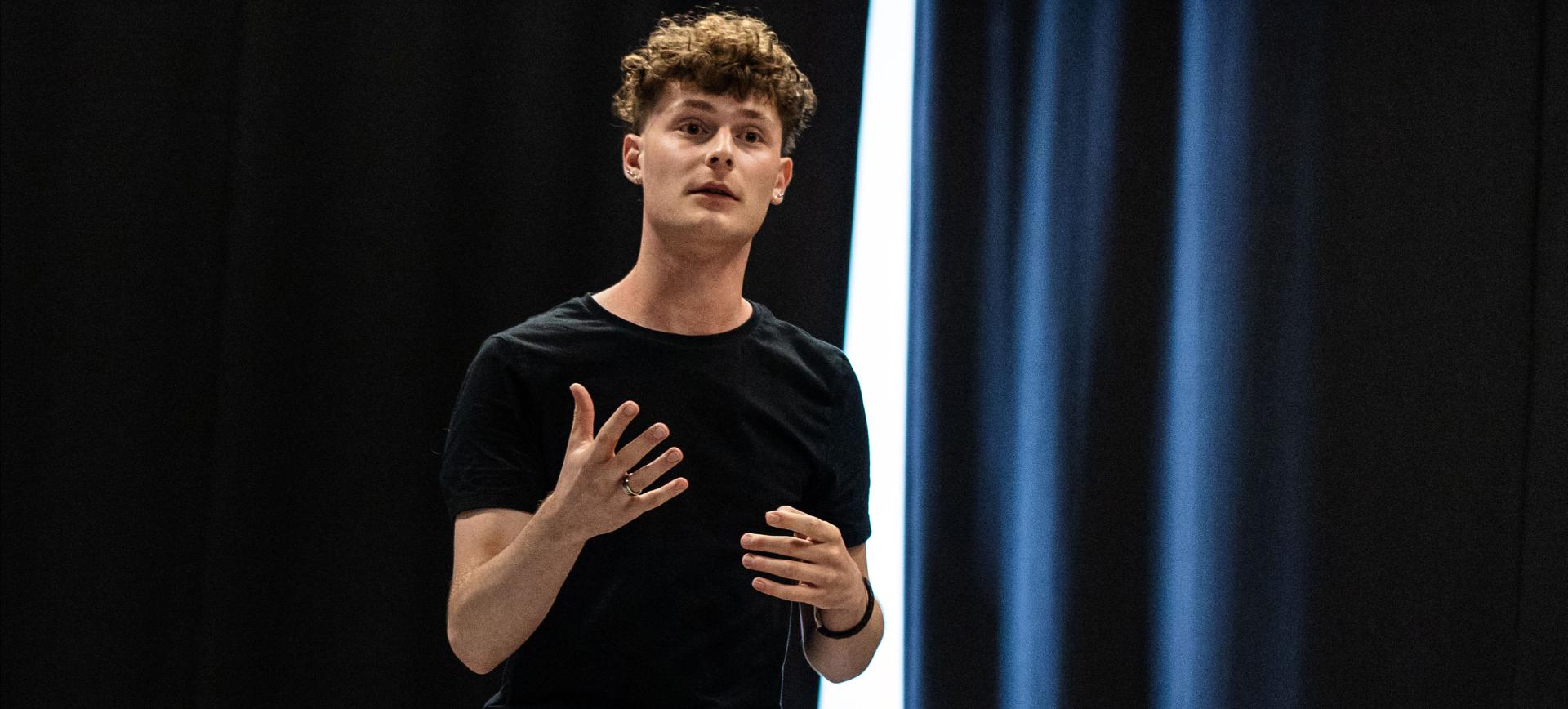 Young man with curly hair speaking on stage, gesturing with both hands