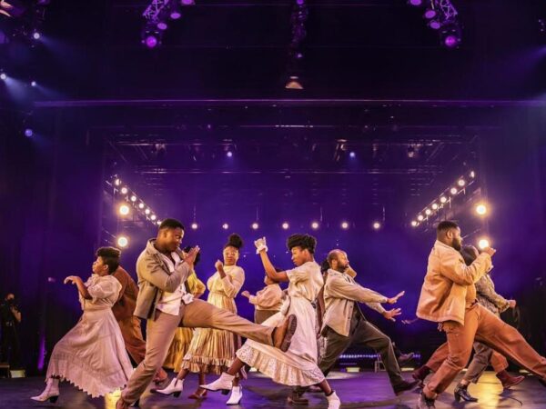 Theatre performers in vintage-style costumes dance energetically on stage under purple lighting