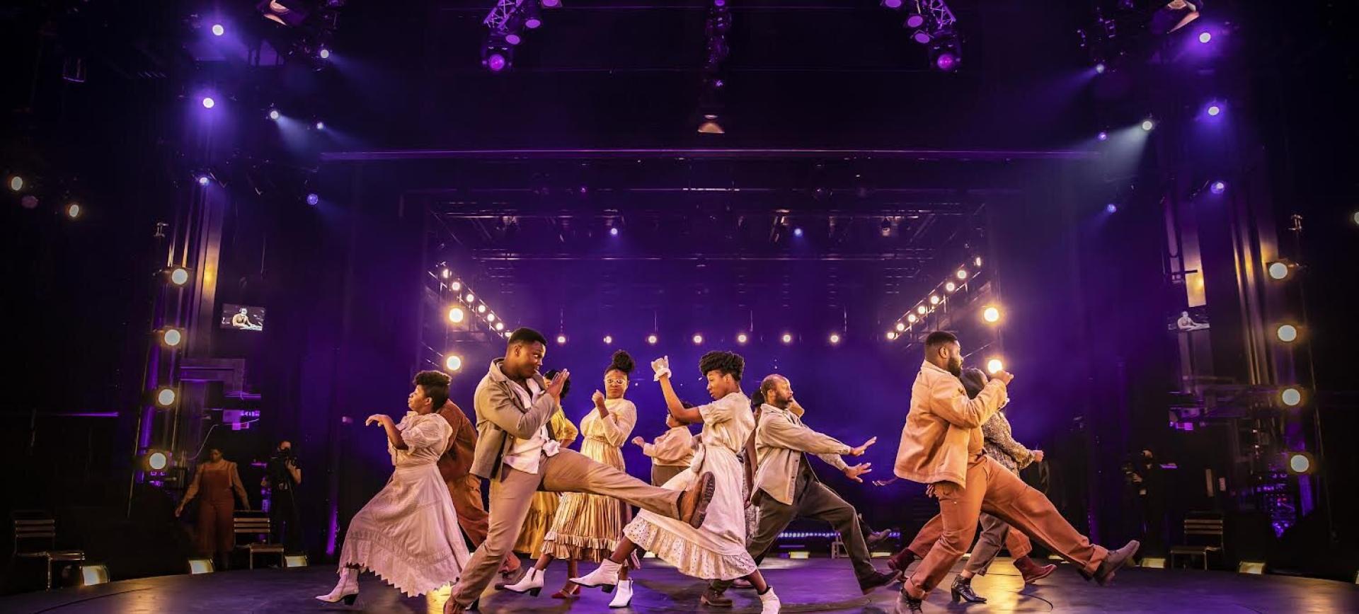 Theatre performers in vintage-style costumes dance energetically on stage under purple lighting