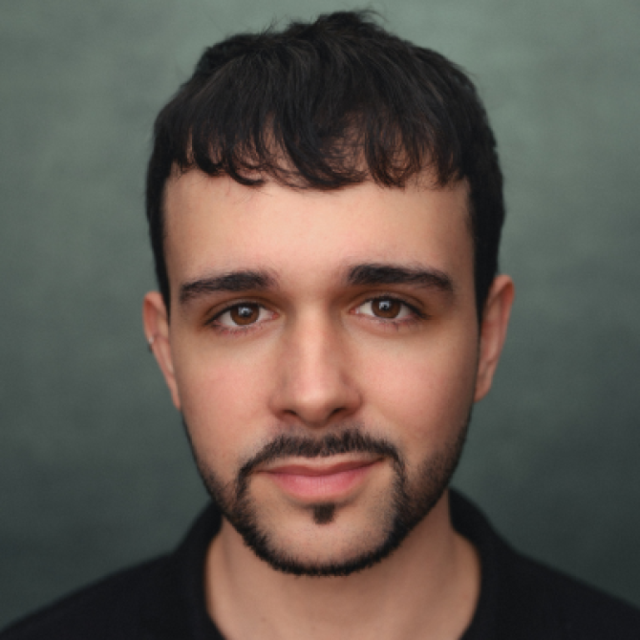 Headshot of a young man with dark hair and a short beard, looking directly at the camera against a plain green-grey background