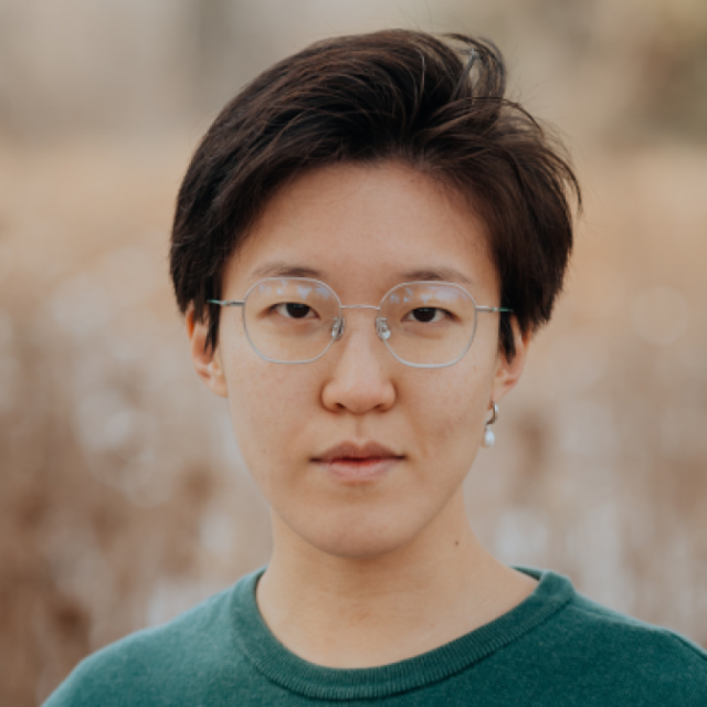 Portrait of a person with round glasses and a pearl earring, wearing a green jumper, outdoors