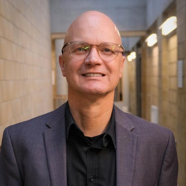 Bald man with glasses, wearing a grey blazer and black shirt, smiling in a corridor