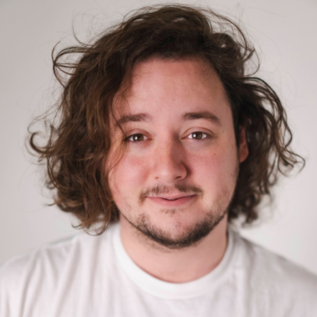 Man with curly shoulder-length brown hair and a short beard, wearing a white T-shirt
