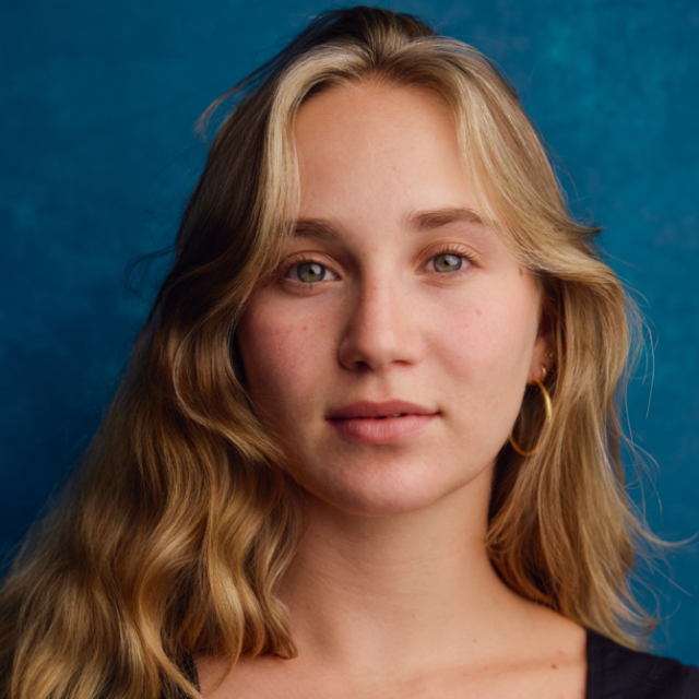 Close-up portrait of a young woman with wavy blonde hair and hoop earrings against a blue background