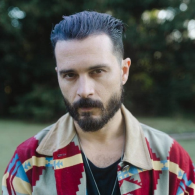 Man with a beard and dark hair, wearing a patterned jacket, standing outdoors in front of trees