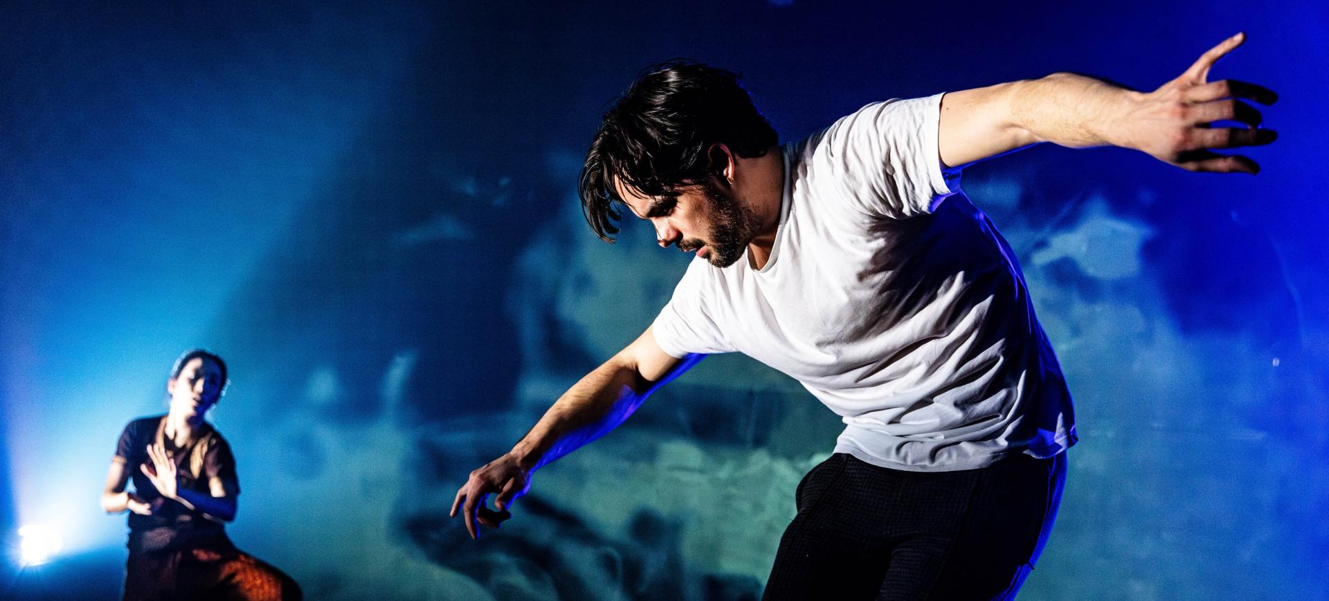 A dancer in a white T-shirt performs an expressive lunging movement under blue stage lighting, with another performer visible in the background
