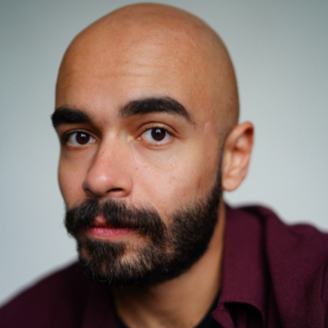Close-up portrait of a bald man with a full dark beard, wearing a maroon shirt