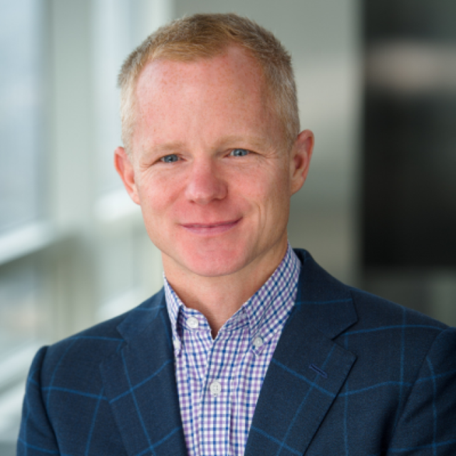 Portrait of a man with short reddish-blond hair, wearing a checked shirt and a navy blazer