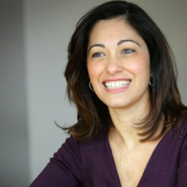 Woman with dark hair smiling, wearing a purple top and a pendant necklace