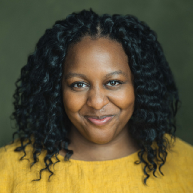 Woman with curly dark hair wearing a mustard yellow top, smiling at the camera against a green background