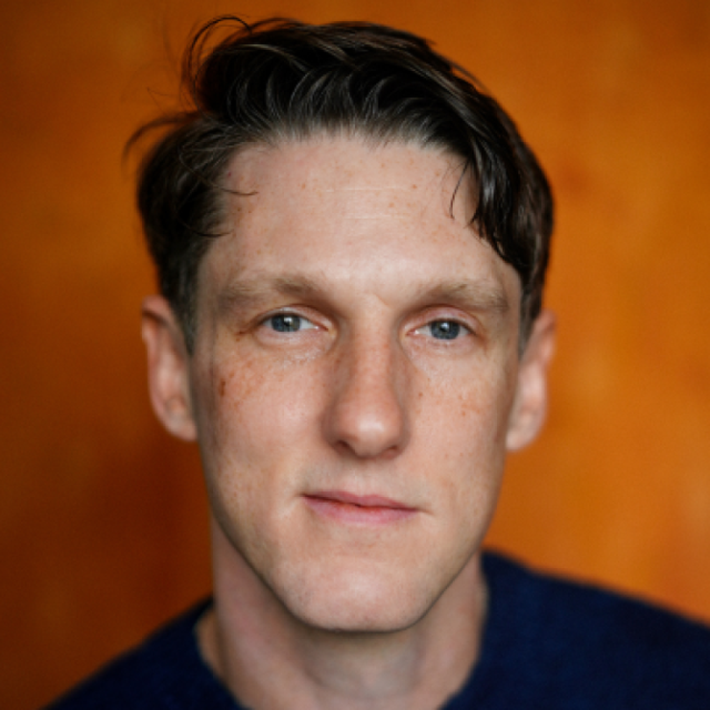 Close-up portrait of a man with dark hair and blue eyes, wearing a navy jumper, against an orange background